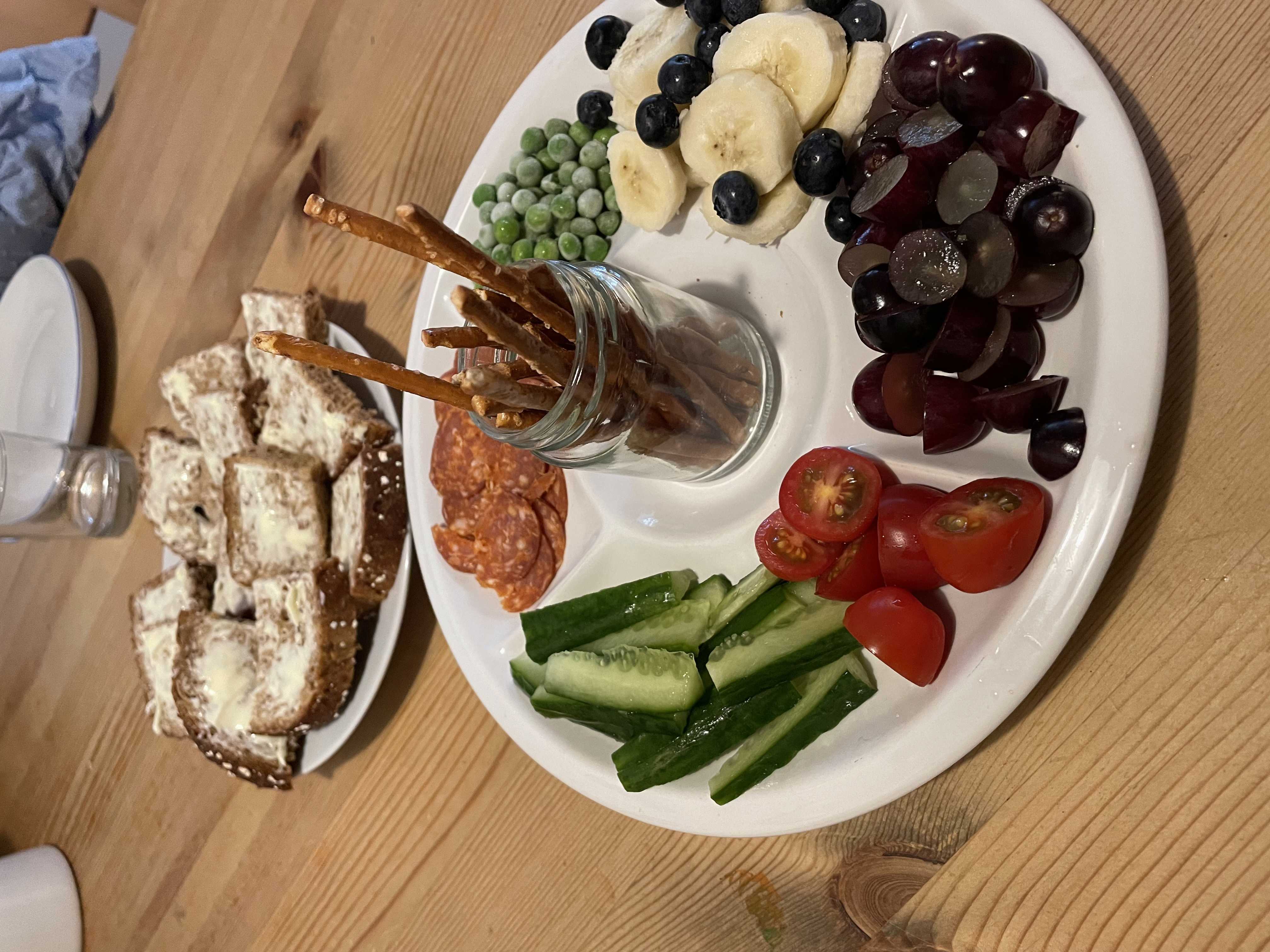 Tallerken med frugt og grønt samt saltstænger. I baggrunden er der en tallerken med grove franskbrødsmadder med smør. 