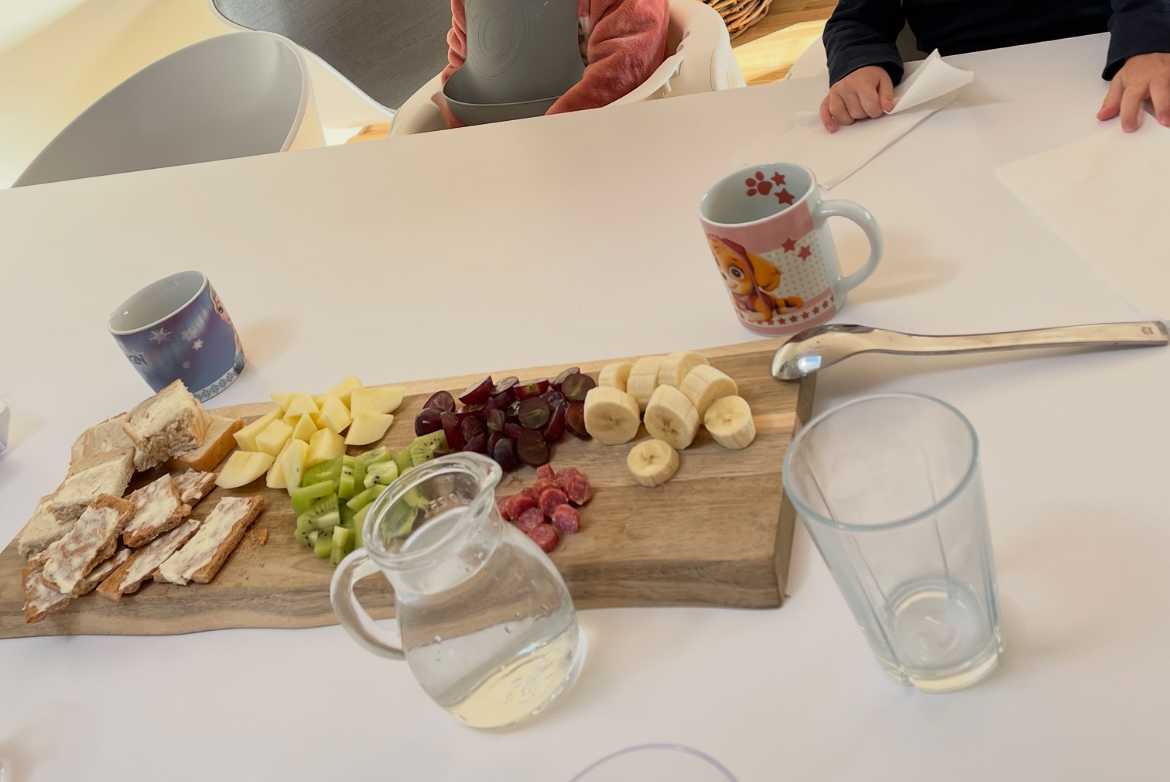 Spækbræt med frugt, brød og spegepølse skåret i mundrette bidder. Der er også vand i kande på bordet, og dagplejebørnene er klar til at spise.