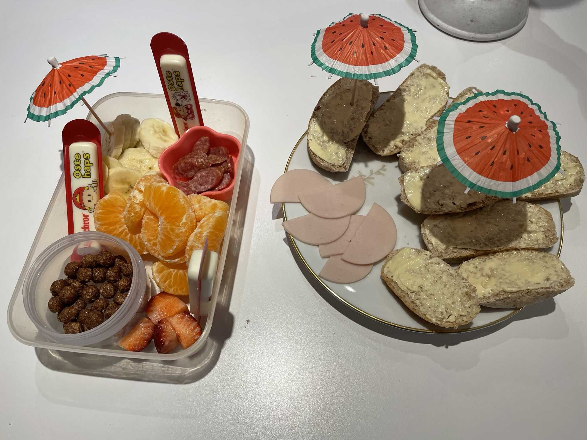 Bøtte med frugt og ostehaps samt tallerken med hjemmebagte boller pyntet med små vandmelonsparasoller.