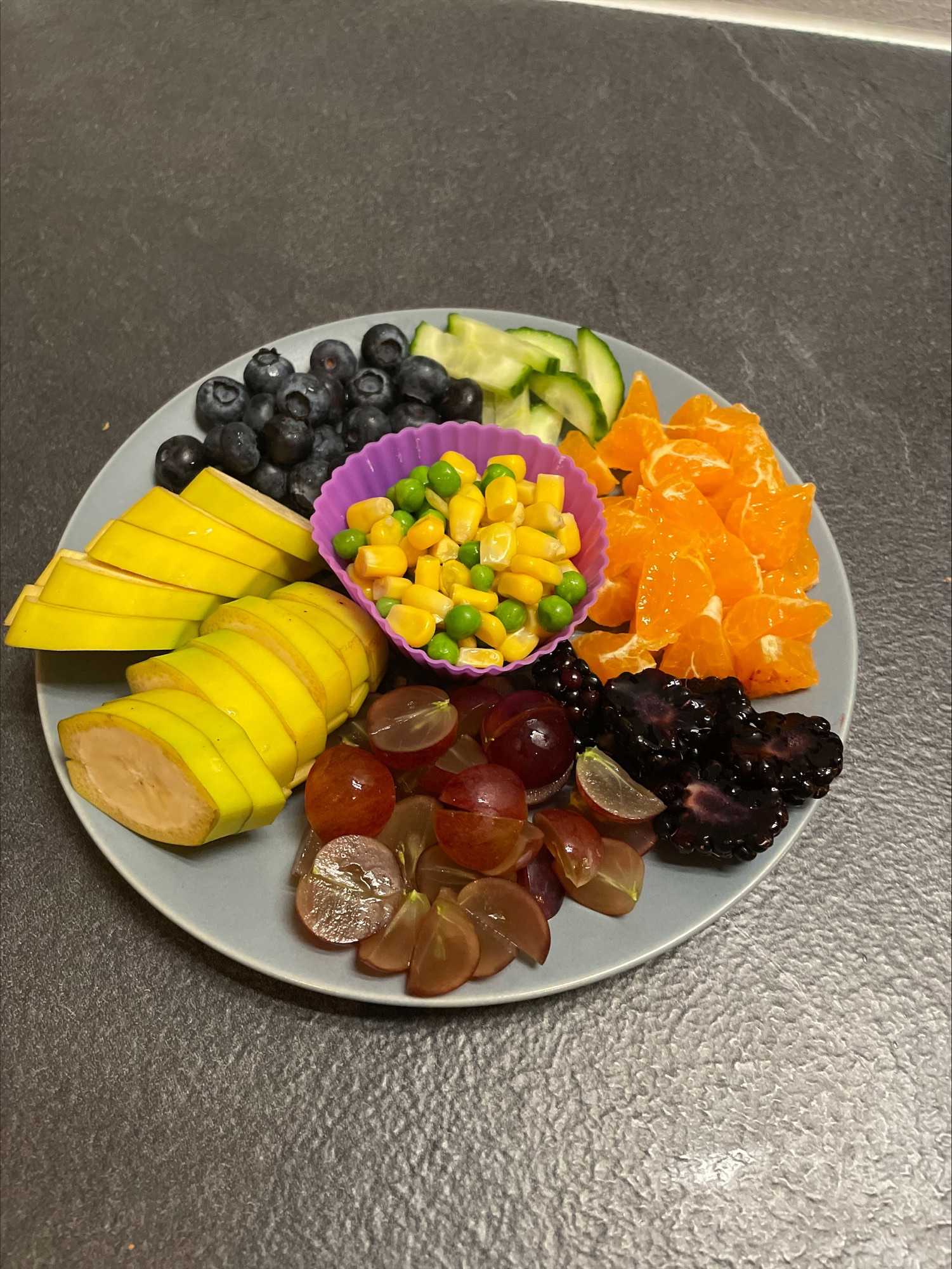 Tallerken med banan, vindrue, clementin, agurker og blåbær skåret i mundrette stykker. Midt på tallerken er der en muffinform med ærter og majs.