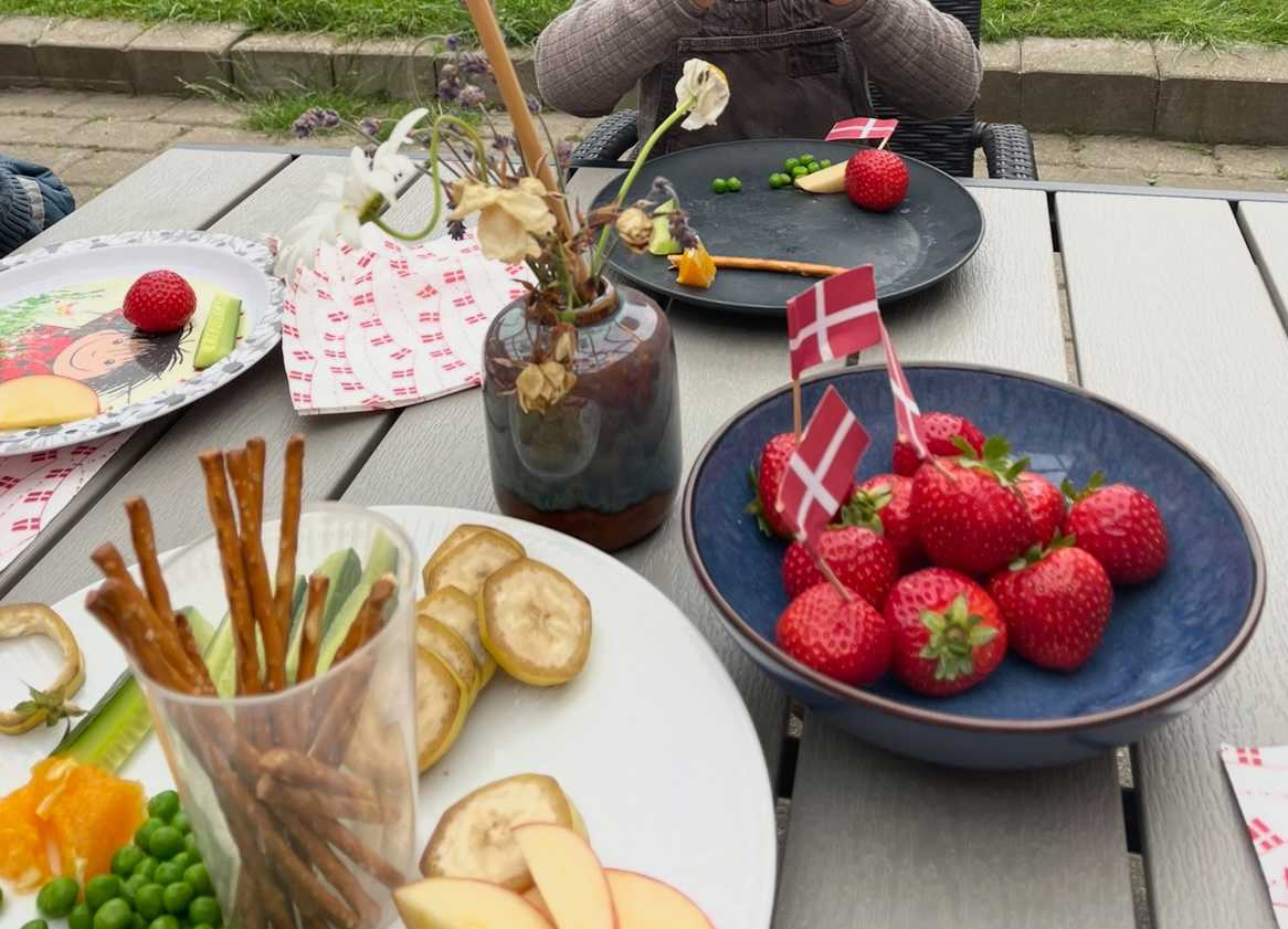 Frokostbord med danske jordbær pyntet med dannebrog, og tallerken med saltstænger, ærter, banan og agurk. Der er en lille buket blomster midt på bordet.
